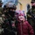 A young girl in a pink cat-themed winter hat and red jacket smiles broadly while posing next to a fully equipped U.S. soldier wearing camouflage, a tactical helmet, balaclava, and carrying a rifle. Additional military personnel in MARPAT (Marine) camouflage uniforms are visible in the background, along with a yellow building that appears to be a church. The scene suggests a public military presence or community engagement event in a cold-weather environment.
