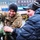 A smiling U.S. Army soldier in MultiCam uniform is interacting with two young civilians, appearing to demonstrate or explain a piece of green military equipment. The soldier wears a unit patch consistent with the 2nd Brigade Combat Team, 1st Armored Division or similar formation, along with a U.S. flag patch. The setting appears to be an outdoor public military display or demonstration in an urban environment during cold weather.
