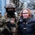 A fully equipped American soldier in tactical combat gear poses with a civilian woman wearing glasses and a dark winter jacket with fur-trimmed hood. The soldier is wearing MultiCam camouflage, a tactical helmet with night vision mount, face covering, and carries a rifle. The setting appears to be an outdoor public event with other military personnel visible in the background.