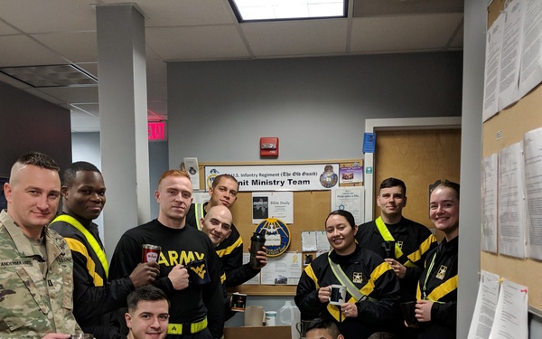 A group of approximately 12 U.S. Army soldiers posing together in what appears to be a Unit Ministry Team office, holding coffee mugs and travel tumblers. A bulletin board in the background displays a sign reading '3rd U.S. Infantry Regiment (The Old Guard) Unit Ministry Team.' The soldiers are a mix of those in Army Combat Uniform (ACU/OCP) and Army Physical Fitness uniforms (black with yellow/gold accents).
