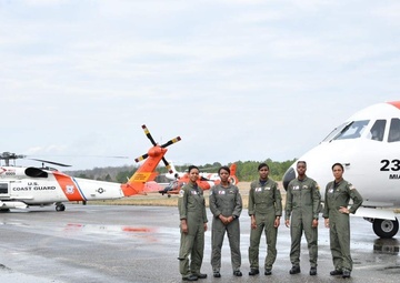 Five U.S. Coast Guard personnel in olive/sage green flight suits are standing on a wet tarmac in front of aircraft. The fixed-wing aircraft on the right is numbered 2318 and marked 'MIAMI,' while a U.S. Coast Guard helicopter (number 6009) is visible on the left. The reflective wet surface suggests recent rainfall at what appears to be a Coast Guard air station.