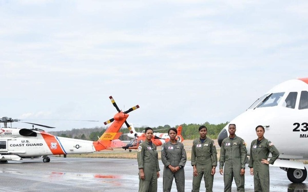 Five U.S. Coast Guard personnel in olive/sage green flight suits are standing on a wet tarmac in front of aircraft. The fixed-wing aircraft on the right is numbered 2318 and marked 'MIAMI,' while a U.S. Coast Guard helicopter (number 6009) is visible on the left. The reflective wet surface suggests recent rainfall at what appears to be a Coast Guard air station.