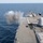 A naval warship is firing its main deck gun, producing a large cloud of white and gray smoke from the muzzle blast. The photograph is taken from an elevated position on the ship looking forward along the bow deck. The vessel appears to be underway on calm, open blue ocean under clear skies.
