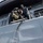 Two military personnel are visible on the deck of a naval vessel, with one individual in camouflage gear and tactical equipment climbing or being assisted over the ship's railing. A sailor in Navy Working Uniform (NWU) blue digital camouflage is assisting the boarding individual. A rope ladder or Jacob's ladder is visible hanging down the gray hull of the ship.