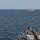 The image shows a naval scene with two warships at sea. The foreground vessel's bow deck is visible, while a second gray warship is observed at a distance on the horizon. The sea conditions appear calm with a clear, hazy sky.