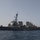 A U.S. Navy Arleigh Burke-class guided-missile destroyer is photographed underway on calm open water under a hazy sky. The vessel is painted in standard naval haze gray and displays typical destroyer armaments and superstructure. Personnel are visible on the deck, suggesting active operations or a transit mission.