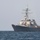 A large gray naval warship is photographed at sea under an overcast sky. The vessel appears to be an Arleigh Burke-class guided-missile destroyer, identifiable by its distinctive hull shape, superstructure, and radar/weapons systems. The ship is underway or stationary on calm, dark blue open water.