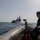 A person in black tactical/dive gear sits on the inflated tube of a rigid inflatable boat (RIB) on open water, observing a naval vessel in the middle distance. The warship, bearing a hull number that appears to be '846', is stationary or moving slowly on calm seas. The scene suggests a maritime military or coast guard operation.