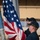 Two U.S. Navy sailors in dark blue uniforms and black ball caps are raising or handling the American flag on what appears to be the deck of a naval vessel. The flag is prominently displayed with its stars and stripes clearly visible. The background shows what appears to be a naval facility or hangar structure.