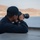 A person dressed in dark navy clothing and a matching baseball cap is photographing from what appears to be the deck of a naval or military vessel. They are using a camera equipped with a long telephoto lens, aimed toward a body of water with arid, rocky hills visible in the background. The setting suggests a maritime surveillance or documentation activity in a coastal region.