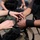 The image shows multiple individuals in military or tactical gear exchanging or handling a roll of black tape in the foreground. The scene appears to be an outdoor staging or preparation area where personnel are gearing up. The focus is tightly on the hands and the tape roll, with other uniformed individuals visible but blurred in the background.