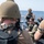 A group of military or law enforcement personnel in tactical gear are gathered on the deck of a naval vessel at sea. The personnel are wearing helmets, body armor, and tactical vests, and appear to be in a briefing or staging posture. The open ocean is visible in the background under clear sky conditions.
