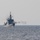 A naval warship is underway at sea, actively firing its deck gun as evidenced by the smoke/muzzle blast visible on the port side. The vessel appears to be a mid-sized military surface combatant with a pennant number visible on the hull (appears to be '731' or similar). The image was taken from a distance under overcast, hazy conditions.