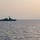 A naval warship bearing the hull number F946 is silhouetted against a hazy sunset sky on calm open waters. The vessel appears to be a frigate or corvette-class warship, photographed at dusk from a distance. A small red buoy or marker is visible on the water to the right of the vessel.