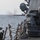 The image shows a naval weapon system mounted on the deck of a warship, actively firing over open ocean. A visible smoke trail at the muzzle indicates the weapon has just discharged. The ship's superstructure, deck hardware, and mooring lines are visible in the foreground and background.