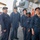 Five U.S. Navy personnel in navy blue coveralls are posing together on the deck of a naval vessel, likely a destroyer based on visible equipment. The group includes both male and female sailors of diverse backgrounds, wearing ship-specific ball caps. They appear to be in port, with dock equipment and structures visible in the background.