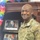 A U.S. Army soldier in OCP (Operational Camouflage Pattern) uniform is posing and smiling in what appears to be a military office. The uniform displays 'U.S. ARMY' tape and a name tape reading 'BEAGLE' along with rank insignia and qualification badges. The background features framed photographs, challenge coins, military memorabilia, and colorful decorative flowers.