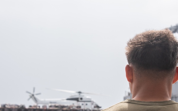 Two individuals wearing olive/dark green t-shirts with Korean Hangul characters and partial text reading 'CAMP M...' are observing a naval vessel from what appears to be another ship or dock. A helicopter is visible hovering or landing on the deck of the ship in the background, with a crowd of people visible on the vessel's deck. The scene suggests a military or naval exercise, possibly involving US Marines at a base in South Korea.