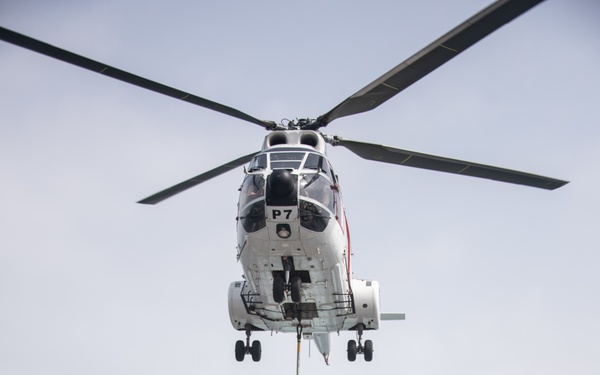 A white military helicopter marked 'P7' hovers over a ship's flight deck, lowering a cargo net filled with supplies via a sling load operation. The helicopter appears to be conducting a vertical replenishment (VERTREP) operation at sea, with the ocean visible in the background. Additional palletized cargo is visible on the deck to the left, suggesting an ongoing resupply mission.