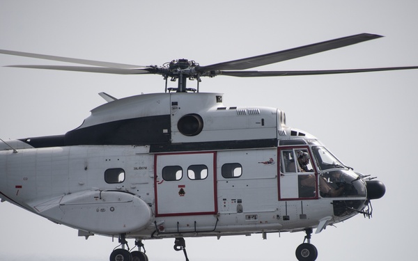 A white civilian helicopter with registration number N814AR is hovering over a ship deck loaded with netted cargo pallets, apparently engaged in a vertical replenishment or cargo transfer operation over open ocean. The helicopter appears to be a Sikorsky S-61 or similar large utility helicopter, identifiable by its twin-engine configuration and distinctive fuselage shape. The operation is taking place in overcast conditions over what appears to be a gray, calm sea.