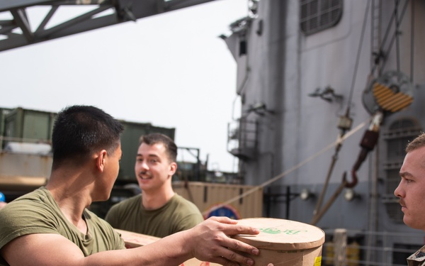 Military personnel, appearing to be U.S. Marines based on their desert MARPAT camouflage uniforms and olive drab t-shirts, are conducting a supply offload or onload operation aboard a naval vessel. They are forming a human chain to pass cylindrical and boxed containers, likely food or supply items, from one person to another. The operation is taking place on the deck of a ship with crane equipment and rigging visible in the background.