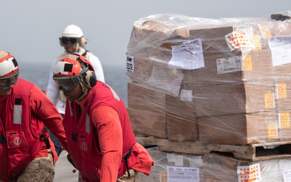 Two personnel in red safety gear and helmets are moving a large palletized cargo load using a red manual pallet jack on what appears to be a ship's deck. The pallet is stacked with cardboard boxes wrapped in clear plastic, bearing labels referencing 'USS Fort McHenry'. A third person in a white helmet is visible in the background near the ocean.