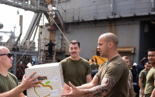 A group of military personnel in U.S. Marine Corps uniforms are conducting a supply transfer operation on the deck of a naval vessel. Two Marines in the foreground are passing a branded cardboard box labeled 'APVLETA' between them, while a line of fellow Marines stands ready in the background. The scene depicts a standard stores or provisions loading evolution aboard a ship.