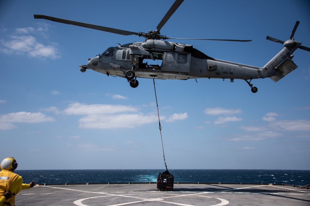 A U.S. Navy MH-60S Seahawk helicopter from HSC-26 is performing a vertical replenishment (VERTREP) operation over the flight deck of a naval vessel at sea. The helicopter, marked with the number 13 and squadron designation HSC-26, is lowering a cargo load via a hoist cable to the ship's deck. A deck crew member in yellow safety gear is visible on the flight deck guiding the operation.