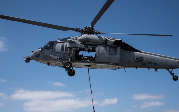 A U.S. Navy MH-60S Seahawk helicopter from HSC-26 is performing a vertical replenishment (VERTREP) operation over the flight deck of a naval vessel at sea. The helicopter, marked with the number 13 and squadron designation HSC-26, is lowering a cargo load via a hoist cable to the ship's deck. A deck crew member in yellow safety gear is visible on the flight deck guiding the operation.