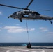A U.S. Navy MH-60S Seahawk helicopter from HSC-26 is performing a vertical replenishment (VERTREP) operation over the flight deck of a naval vessel at sea. The helicopter, marked with the number 13 and squadron designation HSC-26, is lowering a cargo load via a hoist cable to the ship's deck. A deck crew member in yellow safety gear is visible on the flight deck guiding the operation.
