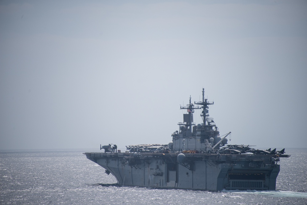 A large amphibious assault ship is underway at sea under overcast, hazy conditions. The vessel has a flat flight deck loaded with multiple aircraft, including what appear to be fixed-wing jets and rotary-wing aircraft. The ship's island superstructure is visible on the starboard side with multiple radar and communication masts.