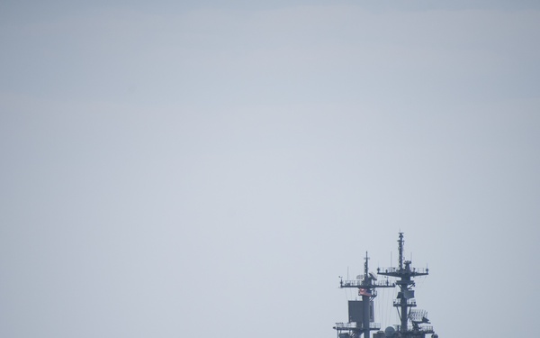 A large amphibious assault ship is underway at sea under overcast, hazy conditions. The vessel has a flat flight deck loaded with multiple aircraft, including what appear to be fixed-wing jets and rotary-wing aircraft. The ship's island superstructure is visible on the starboard side with multiple radar and communication masts.