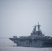 A large amphibious assault ship is underway at sea under overcast, hazy conditions. The vessel has a flat flight deck loaded with multiple aircraft, including what appear to be fixed-wing jets and rotary-wing aircraft. The ship's island superstructure is visible on the starboard side with multiple radar and communication masts.