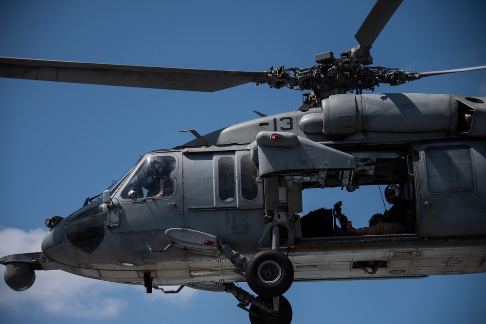 A military helicopter, identifiable as a Sikorsky UH-60 Black Hawk or similar variant, is captured in flight against a clear blue sky. The aircraft bears the number '13' on its fuselage and is painted in standard military gray. At least two crew members are visible — one in the cockpit and one in the open side door.