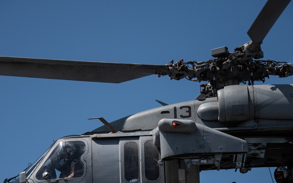 A military helicopter, identifiable as a Sikorsky UH-60 Black Hawk or similar variant, is captured in flight against a clear blue sky. The aircraft bears the number '13' on its fuselage and is painted in standard military gray. At least two crew members are visible — one in the cockpit and one in the open side door.