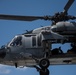 A military helicopter, identifiable as a Sikorsky UH-60 Black Hawk or similar variant, is captured in flight against a clear blue sky. The aircraft bears the number '13' on its fuselage and is painted in standard military gray. At least two crew members are visible — one in the cockpit and one in the open side door.