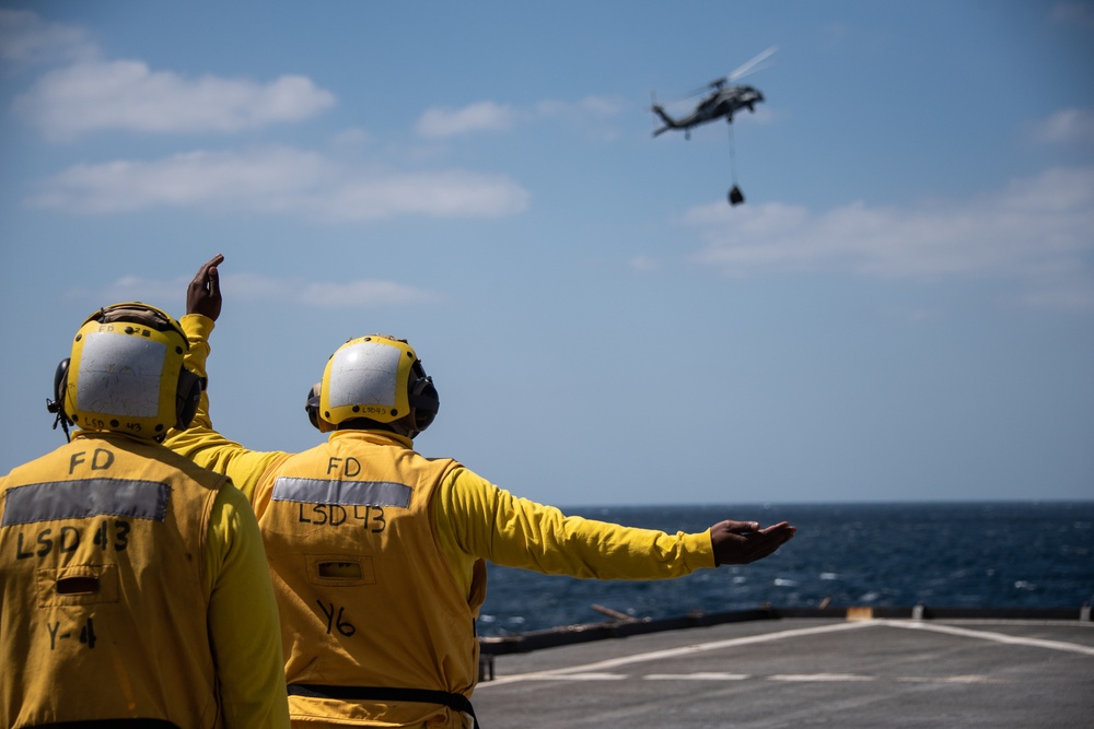 Two naval flight deck personnel wearing yellow 'FD' (Flight Deck) jerseys marked 'LSD 43' are directing a helicopter from the flight deck of USS Fort Fisher (LSD-43). The helicopter, visible in the upper right, appears to be an MH-60 Seahawk conducting a vertical replenishment (VERTREP) operation with a cargo load suspended beneath it. The scene takes place over open ocean under partly cloudy skies.