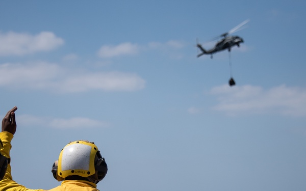 Two naval flight deck personnel wearing yellow 'FD' (Flight Deck) jerseys marked 'LSD 43' are directing a helicopter from the flight deck of USS Fort Fisher (LSD-43). The helicopter, visible in the upper right, appears to be an MH-60 Seahawk conducting a vertical replenishment (VERTREP) operation with a cargo load suspended beneath it. The scene takes place over open ocean under partly cloudy skies.