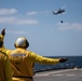 Two naval flight deck personnel wearing yellow 'FD' (Flight Deck) jerseys marked 'LSD 43' are directing a helicopter from the flight deck of USS Fort Fisher (LSD-43). The helicopter, visible in the upper right, appears to be an MH-60 Seahawk conducting a vertical replenishment (VERTREP) operation with a cargo load suspended beneath it. The scene takes place over open ocean under partly cloudy skies.