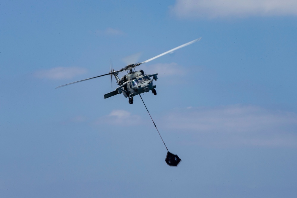 A military helicopter, consistent with an MH-60 Seahawk variant, is captured in flight against a partly cloudy blue sky. The helicopter is towing or deploying a dark object suspended on a long cable or line beneath it. The rotor blades show motion blur, indicating active flight.