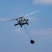 A military helicopter, consistent with an MH-60 Seahawk variant, is captured in flight against a partly cloudy blue sky. The helicopter is towing or deploying a dark object suspended on a long cable or line beneath it. The rotor blades show motion blur, indicating active flight.