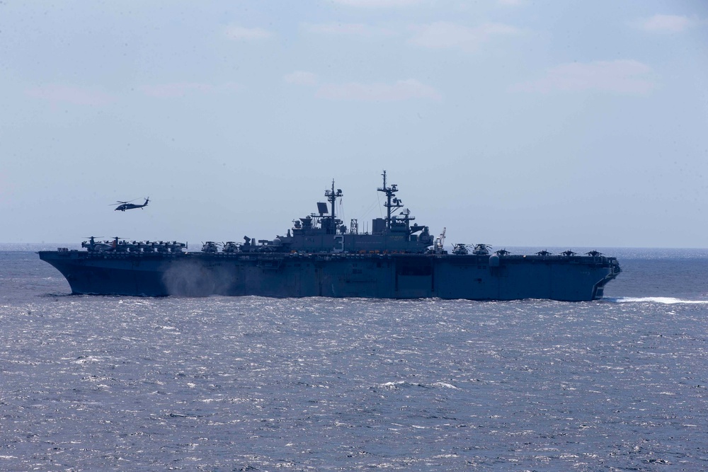 A large US Navy amphibious assault ship is underway at sea, viewed from a distance at a broadside angle. Multiple rotary-wing aircraft are visible on the flight deck and one helicopter is airborne to the port side of the vessel. The ship appears to be conducting flight operations in open ocean conditions.