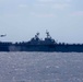 A large US Navy amphibious assault ship is underway at sea, viewed from a distance at a broadside angle. Multiple rotary-wing aircraft are visible on the flight deck and one helicopter is airborne to the port side of the vessel. The ship appears to be conducting flight operations in open ocean conditions.