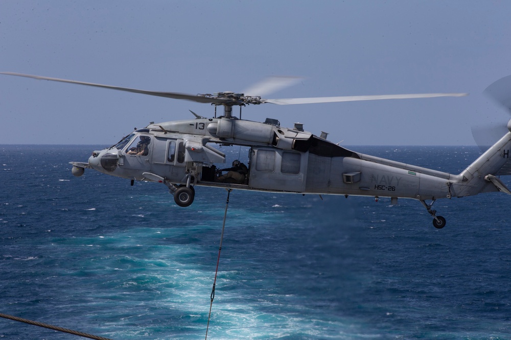 A U.S. Navy MH-60S Seahawk helicopter from Helicopter Sea Combat Squadron 26 (HSC-26), marked with the number 13, hovers over open ocean. The helicopter has its side door open with crew members visible, and a rope or cable is suspended beneath it, suggesting a fast-rope or hoist operation in progress. The image was taken from an adjacent vessel or platform over what appears to be a deep blue ocean.