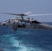 A U.S. Navy MH-60S Seahawk helicopter from Helicopter Sea Combat Squadron 26 (HSC-26), marked with the number 13, hovers over open ocean. The helicopter has its side door open with crew members visible, and a rope or cable is suspended beneath it, suggesting a fast-rope or hoist operation in progress. The image was taken from an adjacent vessel or platform over what appears to be a deep blue ocean.