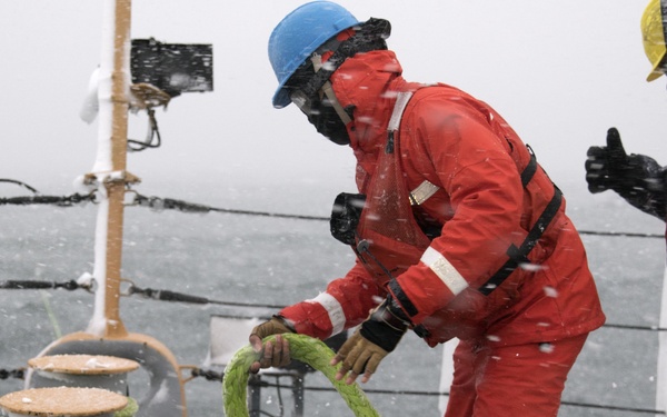 Two workers are operating on the deck of a vessel in severe winter weather conditions with snow and ice visible. The primary worker, dressed in a red survival suit and blue hard hat, is handling a green mooring rope near a deck bollard. The scene depicts challenging maritime deck operations in freezing conditions.