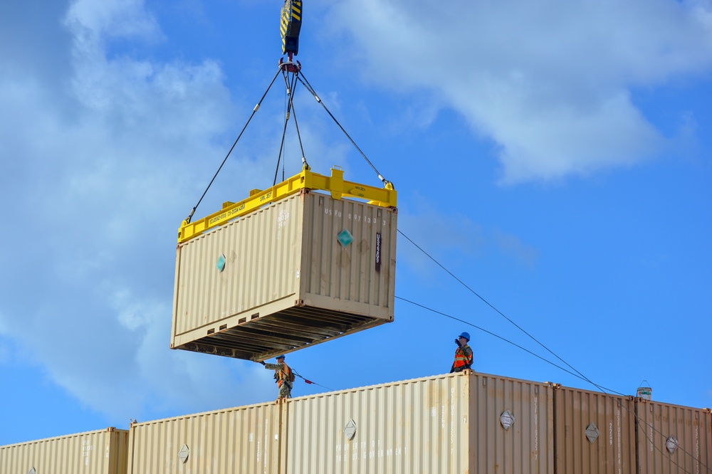 A crane is lifting a beige/tan intermodal shipping container using a yellow spreader bar with wire rigging cables. Two workers are visible standing on top of stacked shipping containers below, guiding the suspended container during placement. The operation appears to be taking place at a port or logistics yard under partly cloudy skies.