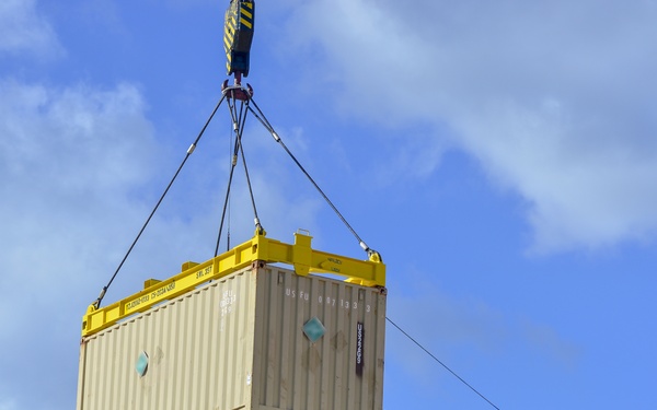 A crane is lifting a beige/tan intermodal shipping container using a yellow spreader bar with wire rigging cables. Two workers are visible standing on top of stacked shipping containers below, guiding the suspended container during placement. The operation appears to be taking place at a port or logistics yard under partly cloudy skies.
