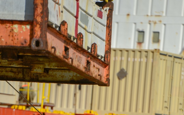 A military service member wearing US Navy digital camouflage (NWU Type III), an orange high-visibility safety vest, blue hard hat, and black gloves is guiding a suspended cargo load using a tag line at a port or dock facility. The individual appears to be acting as a rigger or line handler, controlling the swing of a large rusted metal platform or cargo frame being lifted by a crane. The background shows a ship's hull, shipping containers, and various port infrastructure.