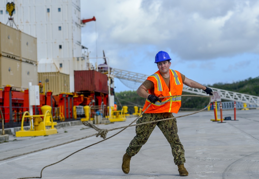 A worker in military-style camouflage pants and a high-visibility orange safety vest is pulling a thick mooring rope on a port or naval pier. He is wearing a blue hard hat and black gloves, indicating active dock work near a large vessel. The setting appears to be a military or commercial port facility with shipping containers and dock equipment visible in the background.